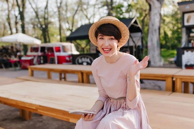 Une fille brune excitée au chapeau à la mode a reçu un message d'un ami et riait. Portrait d'une jeune femme joyeuse en robe vintage, assise avec un téléphone et agitant sa main avec une expression de visage heureux.