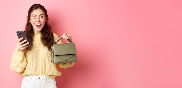 Une fille brune excitée a commandé un sac à main dans une boutique en ligne tenant un smartphone montrant son nouveau sac et smili