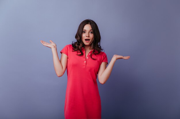 Fille brune extatique aux cheveux ondulés posant avec les mains. Beau modèle féminin européen vêtu d'une élégante robe rouge.