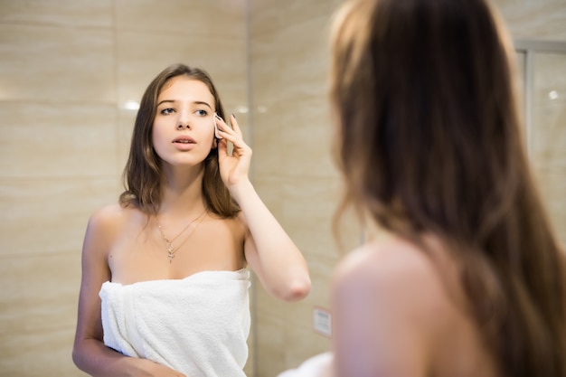 Fille brune fait ses soins de la peau de maquillage du matin dans la salle de bains de mode à la recherche dans le miroir