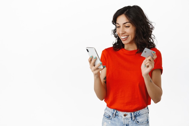 Une fille brune heureuse détient une carte de crédit et a l'air ravie de l'écran du smartphone, vérifie l'application de financement du compte bancaire, achète en ligne, paie avec un téléphone portable, debout sur blanc