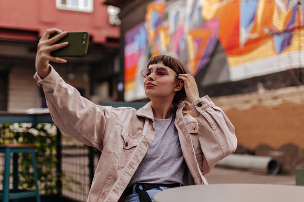 Fille brune hipster faisant selfie à l'extérieur Femme aux cheveux courts en veste beige et lunettes de soleil roses prenant des photos et assis dans un café