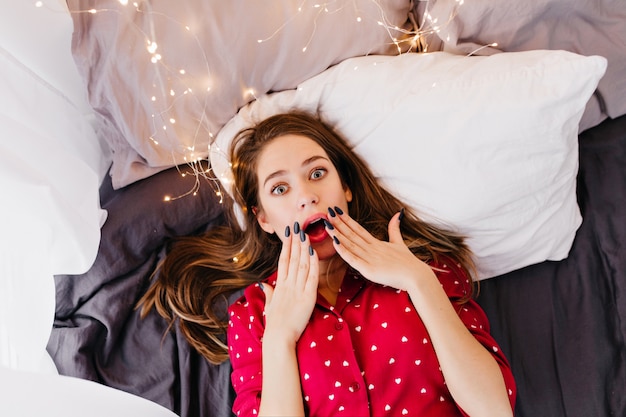 Fille brune inspirée se détendre dans son lit le matin. Portrait intérieur au-dessus de la femme caucasienne rêveuse en pyjama mignon.