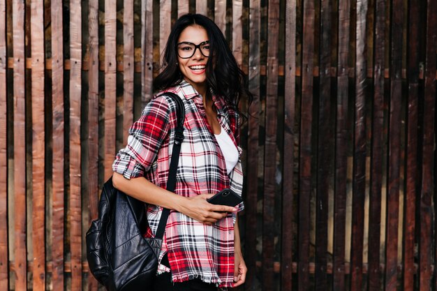 Fille brune inspirée avec téléphone posant sur un mur en bois avec un sourire joyeux. Portrait en plein air d'une femme latine fascinante dans des verres élégants.