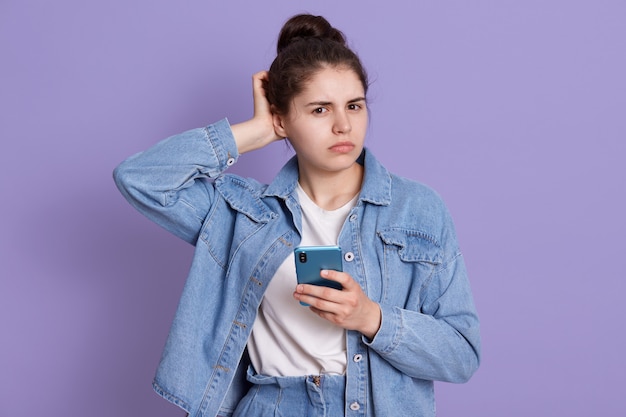 Fille brune perplexe portant une veste en jean et une chemise blanche, gardant la main sur le dos de sa main, tenant un téléphone intelligent