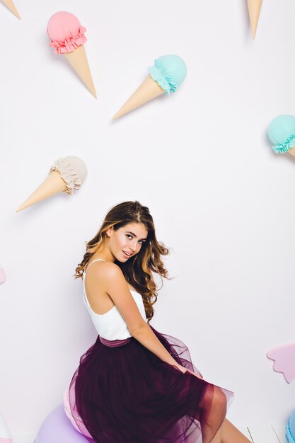 Fille brune romantique en jupe longue violette à la recherche ludique, se reposant dans la chambre avec un intérieur doux. portrait de charmante jeune femme bouclée posant sur le mur décoré avec des trucs de dessert.