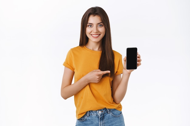 Fille brune séduisante gaie et confiante en t-shirt jaune, tenant un smartphone, pointant un écran mobile et souriant, recommande une application téléphonique géniale, donne un lien vers le code promotionnel, offre un cadeau