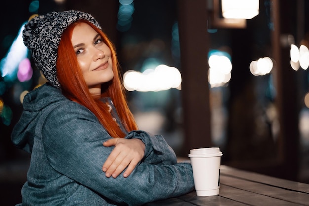 Photo gratuite fille buvant du café chaud assis à la table du marché du soir décoré de lumières de noël. se sentir heureux dans une grande ville. passer les vacances d'hiver