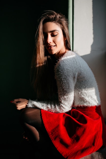 Fille calme aux cheveux longs bruns assis près de la fenêtre et regardant vers le bas