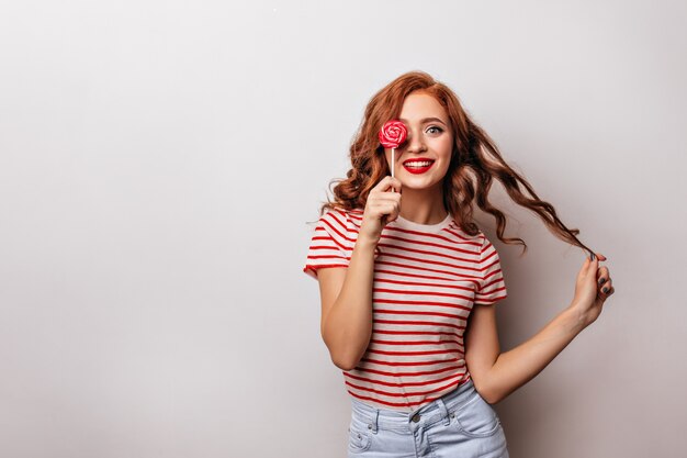 Fille caucasienne intéressée léchant des bonbons avec le sourire. femme au gingembre avec sucette rouge.