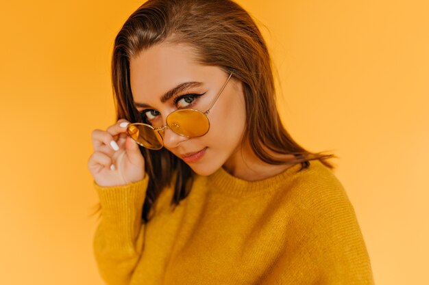 Fille caucasienne à la mode regardant à travers des lunettes de soleil jaunes. Modèle féminin bienheureux en tenue d'automne élégante posant.