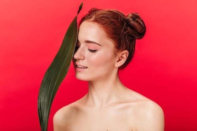 Fille caucasienne rêveuse avec feuille verte souriant sur fond rouge Studio shot of attractive nude gingembre femme avec plante