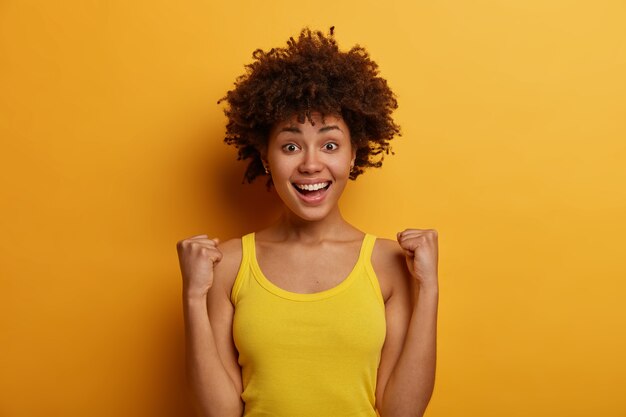 Une fille chanceuse et ambitieuse à la peau sombre fait pomper le poing, célèbre la bonne nouvelle et la réalisation de son objectif, se réjouit de l'excellent événement, porte une chemise jaune décontractée, pose à l'intérieur, rit positivement, sent le triomphe