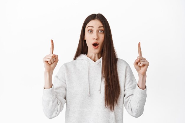 Une fille charismatique impressionnée a l'air fascinée par l'émerveillement devant, la bouche ouverte et haletant étonné, pointant le doigt vers l'annonce promotionnelle cool, mur blanc