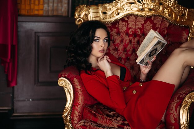Fille chaude en robe rouge courte tenant un livre couché dans la bibliothèque