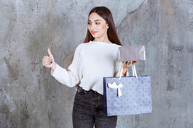 Fille en chemise blanche tenant une boîte-cadeau en argent et un sac à provisions bleu et montrant un signe positif de la main.