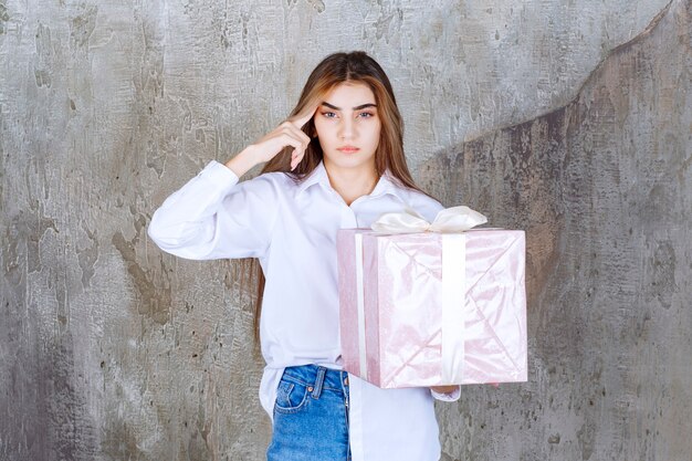 Fille en chemise blanche tenant une boîte-cadeau rose enveloppée d'un ruban blanc et semble confuse et hésitante.