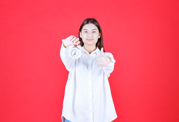 Fille en chemise blanche tenant un réveil et montrant un signe positif de la main