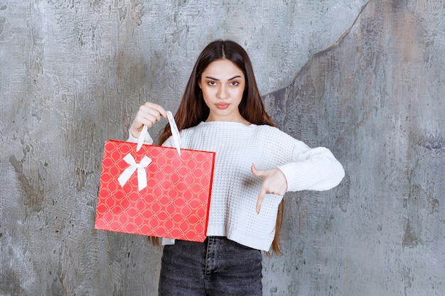 Fille en chemise blanche tenant un sac à provisions rouge et invitant la personne à côté d'elle à présenter le cadeau.