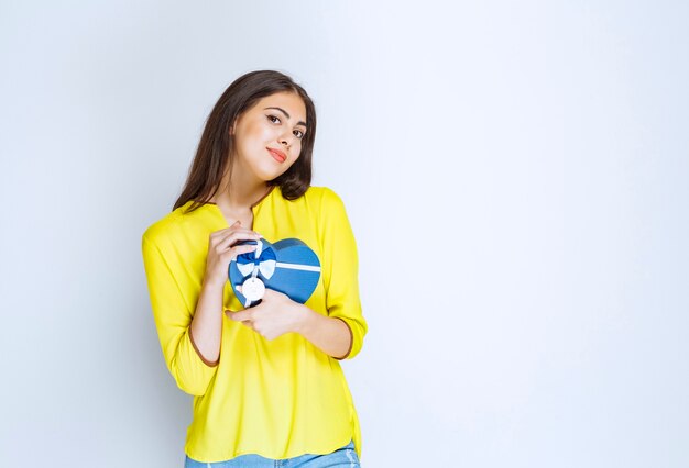 Fille en chemise jaune tenant et promouvant une boîte cadeau en forme de coeur bleu.