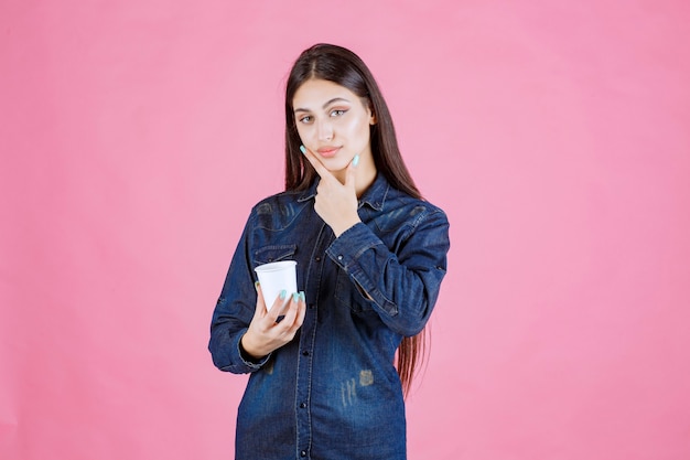 Fille en chemise en jean tenant une tasse de café et a l'air réfléchie et douteuse