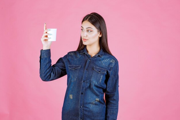 Fille en chemise en jean tenant une tasse de café et a l'air réfléchie et douteuse