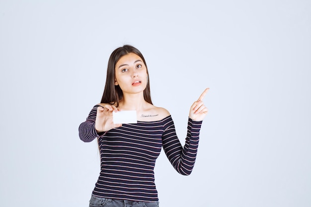 Fille en chemise rayée détient une carte de visite et pointe vers quelqu'un d'autre.