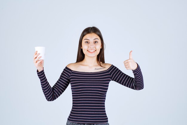 Fille en chemise rayée tenant une tasse à café en plastique et indiquant un bon goût.