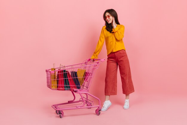 Fille en chemisier orange et lunettes de soleil avec le sourire regarde beaucoup de ses achats se trouvant dans le chariot rose du supermarché.