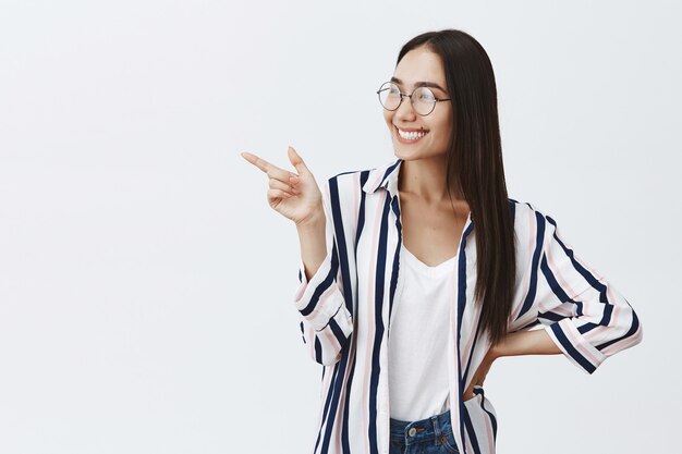 Fille en chemisier tendance rayé et lunettes tenant la main sur la hanche, regardant avec admiration et intérêt, pointant vers la gauche avec l'index en se tenant debout sur un mur gris, assistant à une exposition curieuse