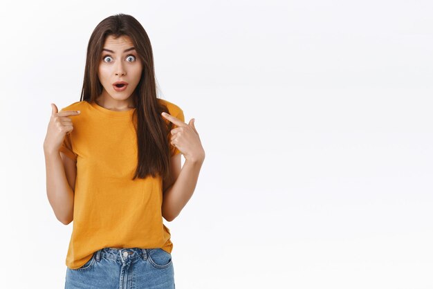 Fille choisie pour le rôle principal dans le jeu debout surprise étonnée se sentant heureuse et étonnée se pointant écarquiller les yeux et souriante reconnaissante avec une expression ravie soulagée fond blanc