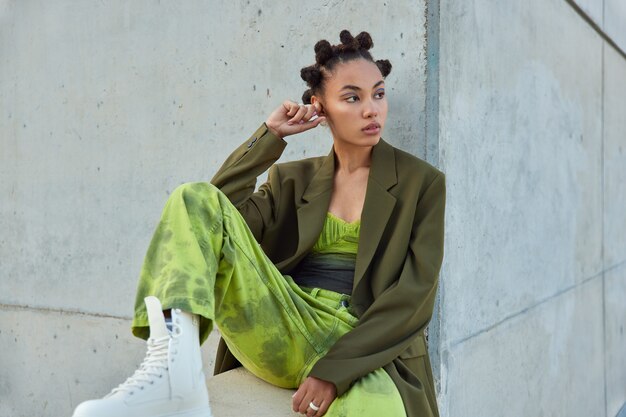 fille avec une coiffure à la mode vêtue de vêtements verts regarde loin pose contre un mur gris urbain considère quelque chose