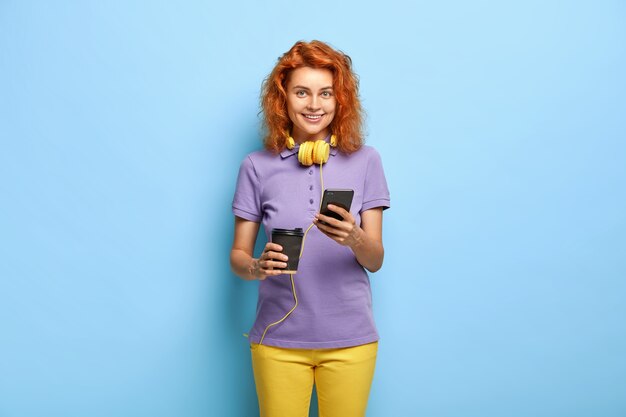 Une fille communicative sociable envoie des messages texte via un téléphone intelligent, boit du café dans une tasse en papier