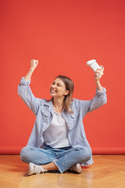 Fille compétitive célèbre gagner tenant le contrôleur de manette de jeu sur mur rouge.