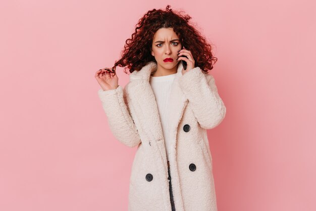 Fille confuse et triste avec des lèvres rouges, parler au téléphone. Portrait de femme en éco-manteau sur espace rose.