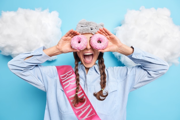 La fille contre les yeux avec des beignets glacés garde la bouche ouverte vêtue d'une chemise avec un ruban s'amuse le jour de l'anniversaire isolé sur bleu