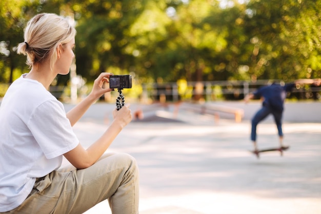 Une fille cool enregistre une nouvelle vidéo d'un jeune patineur sur une planche à roulettes pour un vlog passant du temps au skatepark moderne