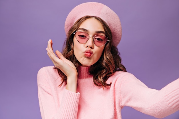 Une fille cool et frisée souffle un baiser et prend un selfie sur un mur violet