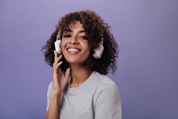 Fille cool en haut gris sourit et écoute de la musique sur un mur violet