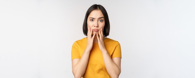 Fille coréenne choquée touchant son visage regardant la caméra inquiète debout en t-shirt jaune sur fond blanc