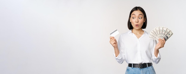 Fille coréenne excitée regardant une carte de crédit détenant de l'argent en espèces posant sur fond de studio