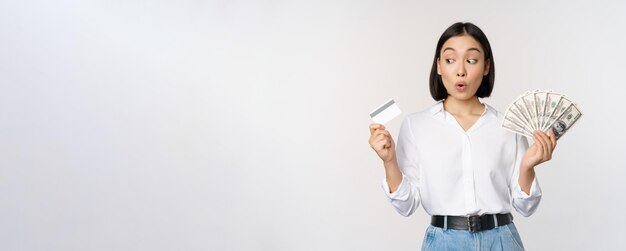 Fille coréenne excitée regardant une carte de crédit détenant de l'argent en espèces posant sur fond de studio