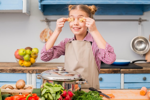 Fille couvrant ses yeux avec des pâtes farfalle debout dans la cuisine