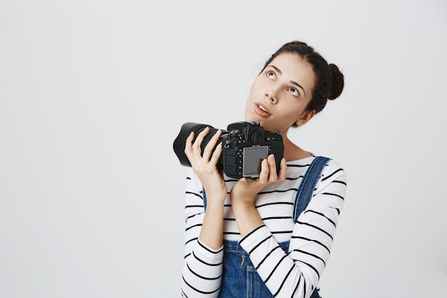 Fille créative réfléchie tenant une caméra professionnelle et regardant dans le coin supérieur gauche, en pensant