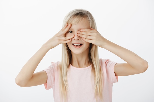 Une fille curieuse veut voir ce que son père a préparé pour le b-day. Portrait de jolie fille impatiente intriguée avec de beaux cheveux blonds, couvrant les yeux avec des paumes et furtivement avec un large sourire sur mur gris