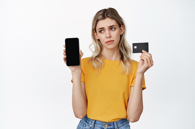 Une fille déçue montre un smartphone vide et une carte de crédit boudant triste debout sur fond blanc