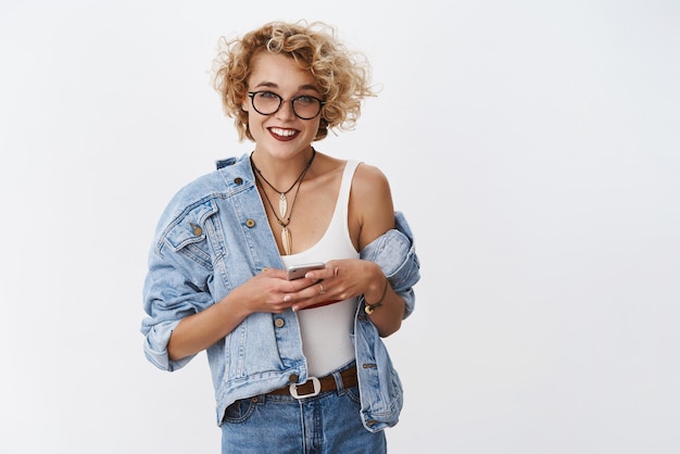 Fille demandant le numéro et écrivant dans le smartphone souriant largement à la caméra amicale posant sans souci dans une pose détendue dans des verres et une veste en jean vérifiant la liste d'épicerie dans le téléphone portable sur fond blanc