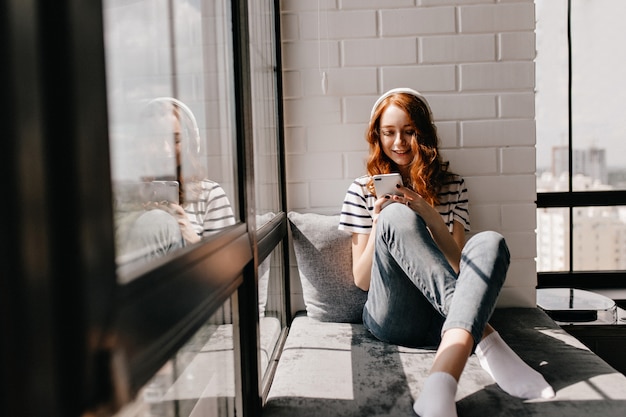 Fille détendue en jeans assis sur le rebord de la fenêtre. Magnifique dame au gingembre dans les écouteurs tenant le téléphone.