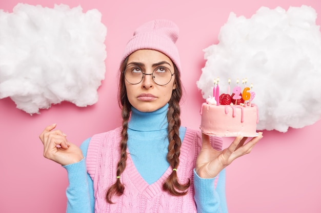 fille avec deux nattes peignées tient un gâteau aux fraises a une expression triste et ennuyée porte un chapeau à col roulé concentré au-dessus isolé sur rose