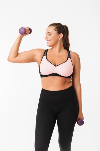 Fille dodue souriante en haut sportif et leggings tenant des haltères dans les mains tout en regardant joyeusement de côté sur fond blanc